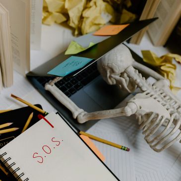 Skeleton slumped on laptop with S.O.S. note amid crumpled papers and sticky notes.