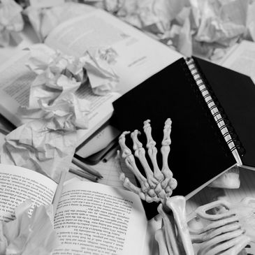 Skeleton hand rests among open books and crumpled papers, symbolizing study stress.