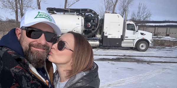 Man and woman in front of white hydrovac truck with snow on ground. Woman kissing man.