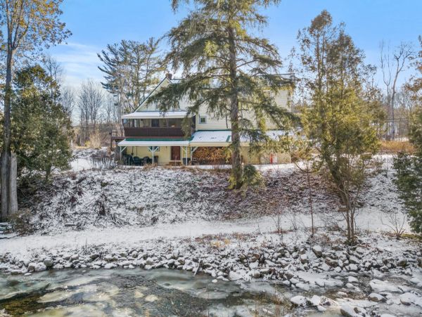 A snowy house near a small stream with trees around it.
