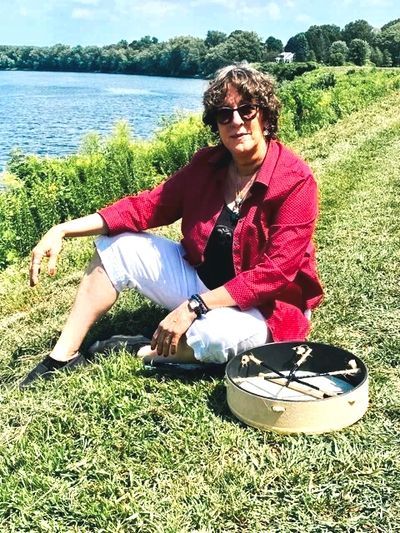 Woman in sunglasses sitting by a river with a drum on the grass.