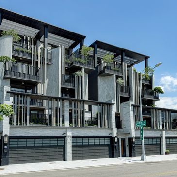Modern multi-story residential building with balconies and greenery under a clear blue sky.