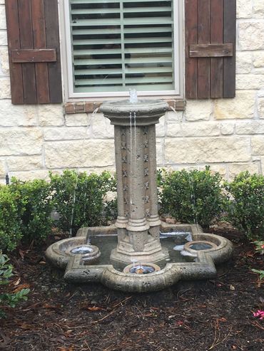 Decorative stone water fountain in garden bed by window with wooden shutters.
