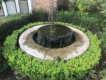 Modern, circular stone water fountain surrounded by green bushes in a garden.
