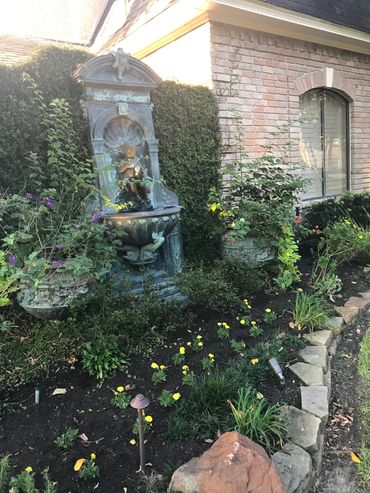 A decorative garden fountain surrounded by flowers and greenery beside a brick house.