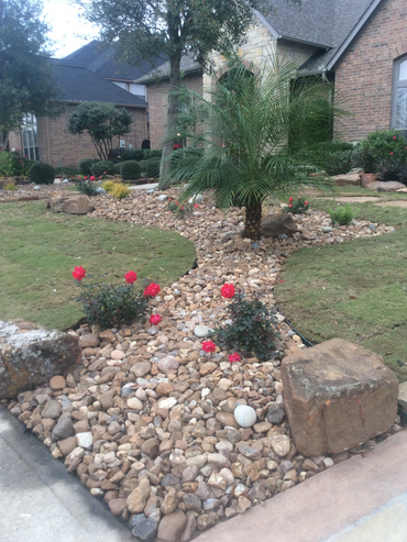 A landscaped front yard with rocks, palm tree, and red flowers near a brick house.