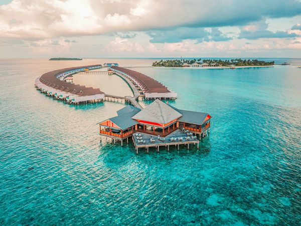 Overwater bungalows and a central pavilion on clear tropical waters under a partly cloudy sky.