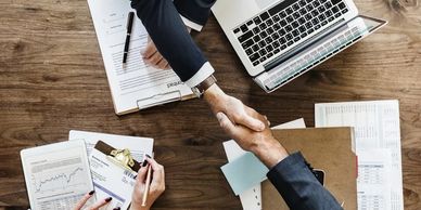 Two people shaking hands over a desk with documents and a laptop.