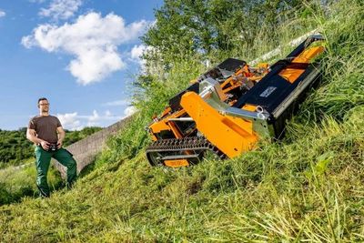 Man operating an orange remote-controlled slope mower on a grassy hill.