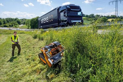 A compact robotic mower cutting tall grass on a roadside slope.