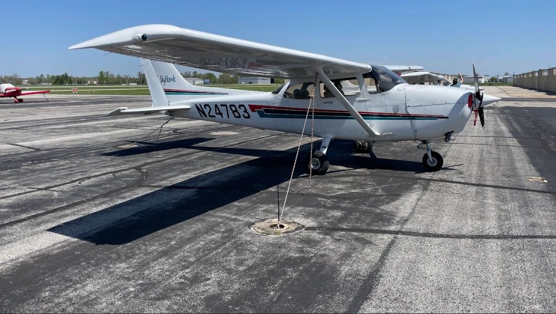 A white Cessna 172 (N24783) with blue and red stripes parked on an asphalt ramp under a clear sky.