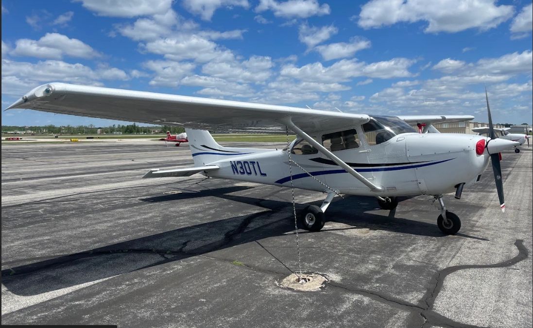 A white Cessna 172 (N30TL) with blue stripes parked on an asphalt ramp under a partly cloudy sky.