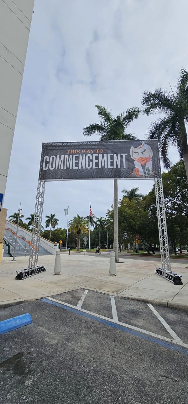Sign directing to commencement with palm trees and flags in background.