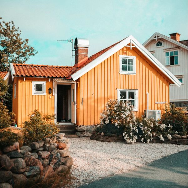 Cozy yellow house with white trim surrounded by a garden and gravel driveway.