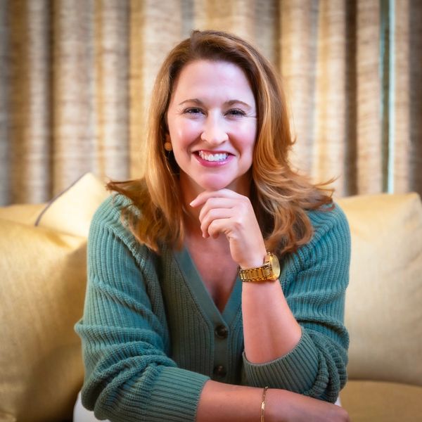 Smiling woman in green sweater sitting on a couch with hand on chin.