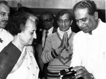 Black and white photo of a woman in a sari and men in formal attire, one man showing something to the woman.