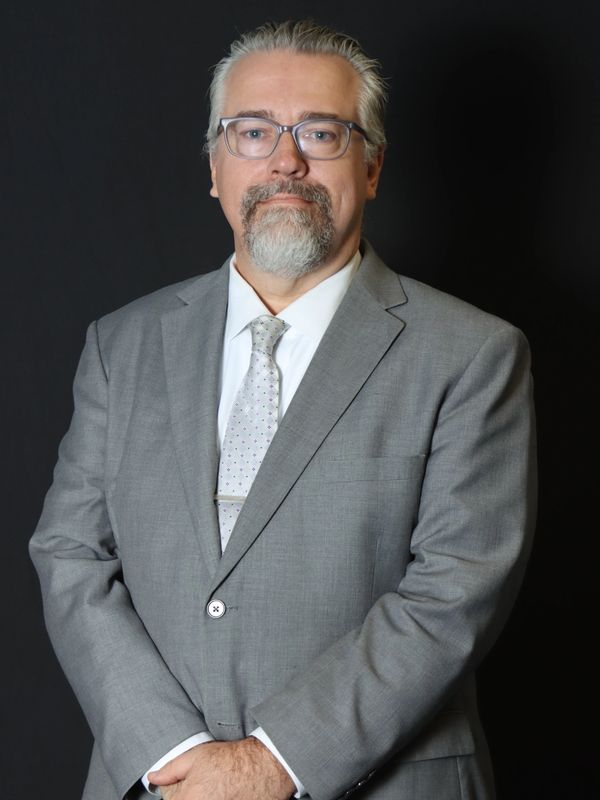 Middle-aged man in a gray suit and patterned tie standing against a black background.