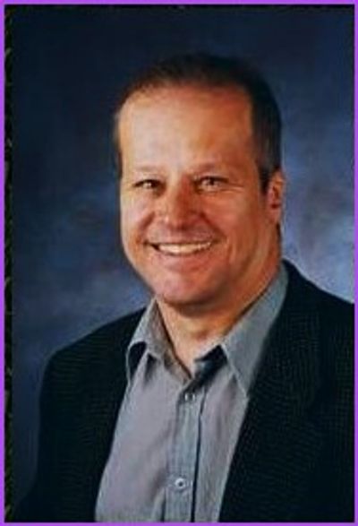 Smiling man in a suit jacket with a grey shirt against a dark background.