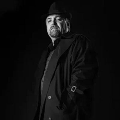 Man in a dark coat and hat posing confidently against a black background.