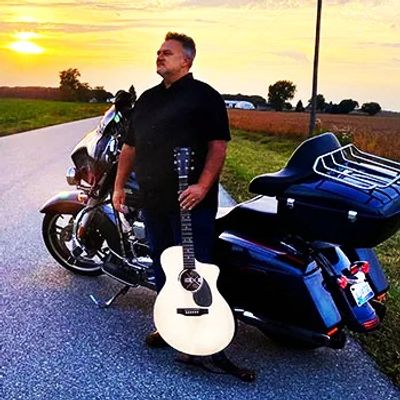 Man stands by motorcycle holding guitar at sunset on a rural road.