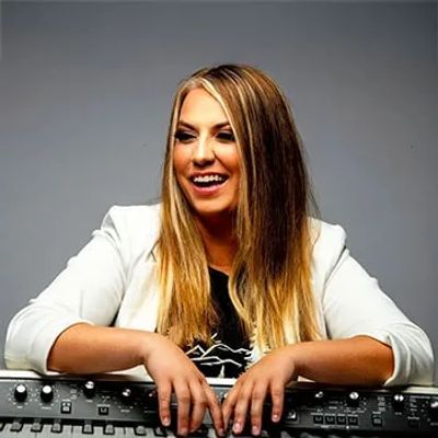 Smiling woman in a white blazer with hands resting on audio equipment.