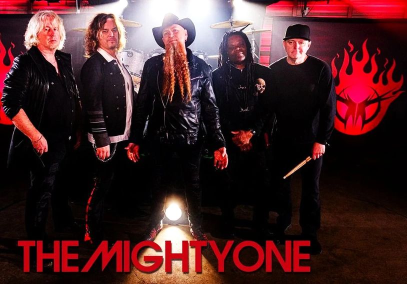 The Mighty One band posing with instruments and logo in background.