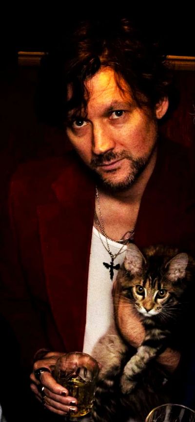 Man in red jacket holding a cat and a glass of drink.