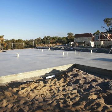 Concrete foundation slab set on a sandy construction site under clear blue sky.