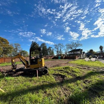 A small excavator works on a sunny, partly cloudy day in a grassy field near houses.