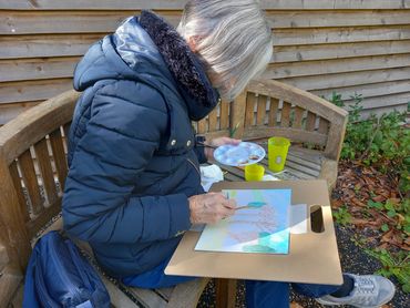 Elderly woman painting outdoors on a wooden bench.