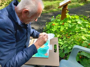 Elderly man painting outdoors beside green plants under sunlight.