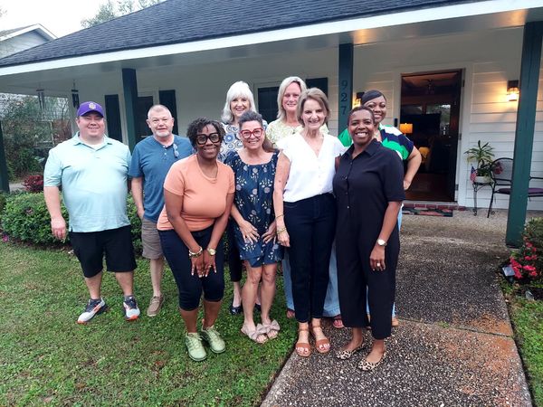 A group of eight adults smiling in front of a house.