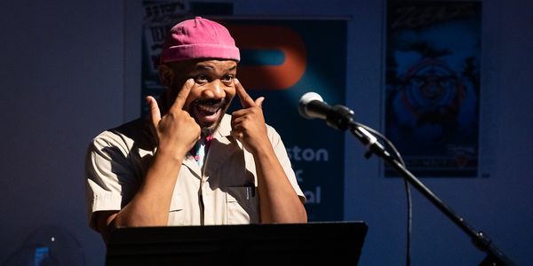 Man speaking at a microphone, gesturing with fingers near his eyes.