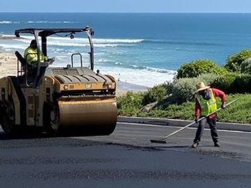 Road workers paving asphalt near a coastal area with ocean waves.