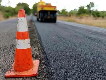 Orange traffic cone on a freshly paved road with a road roller in the background.