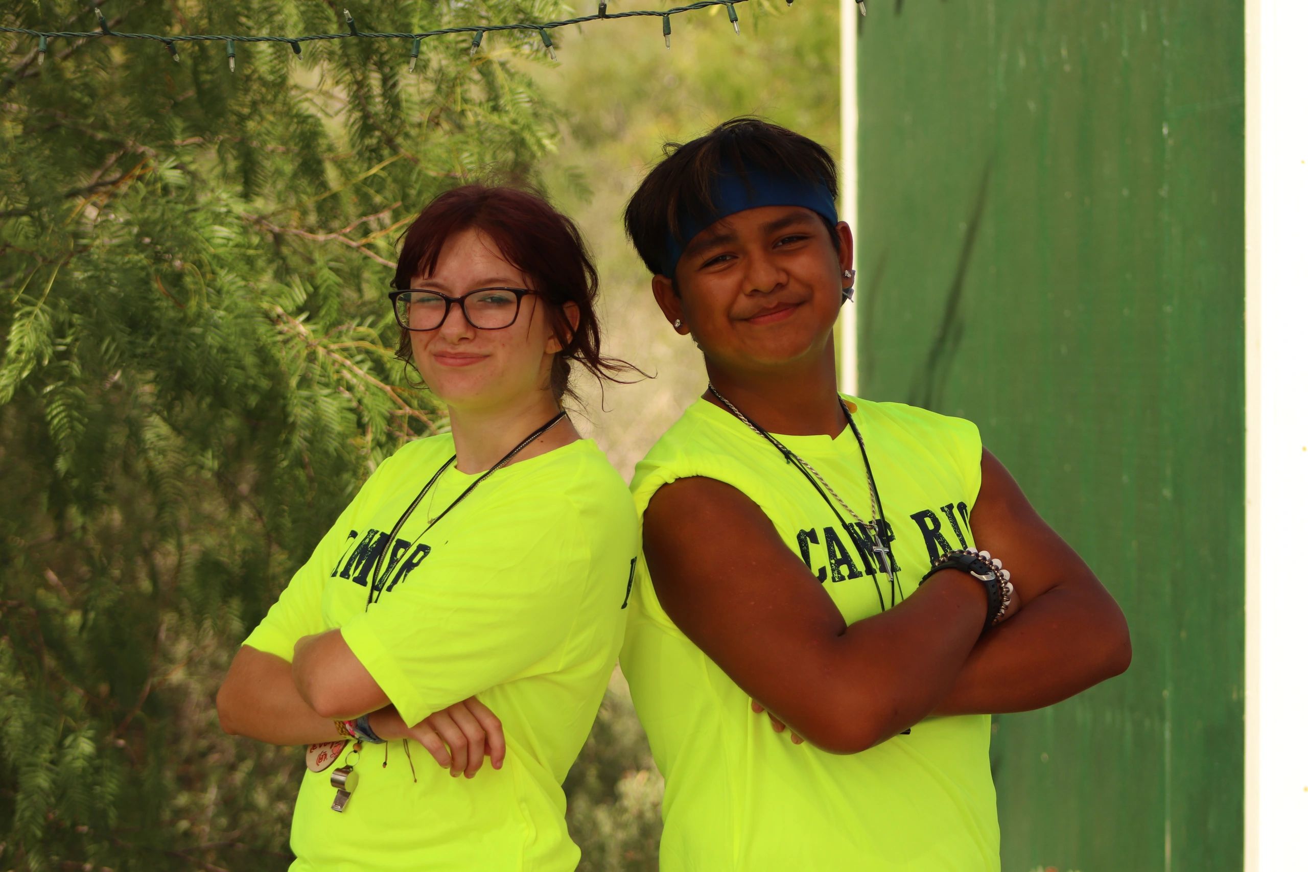 Two smiling teens in bright yellow shirts pose back to back outdoors.