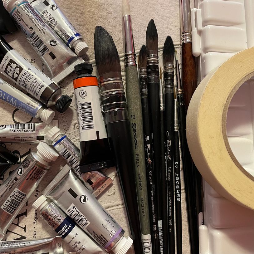 Various paintbrushes and tubes of paint arranged on a surface with masking tape.