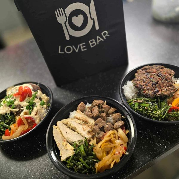 Three bowls of diverse meals in front of a black Love Bar takeout box.