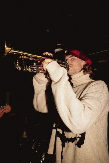 Man in a red hat playing trumpet with eyes closed in a dark setting.