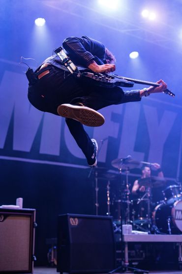 Guitarist jumps high on stage during a live rock concert with drummer in background.
