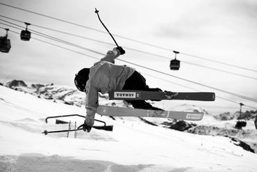 Skier performing an aerial trick on snowy slopes with cable cars in the background.