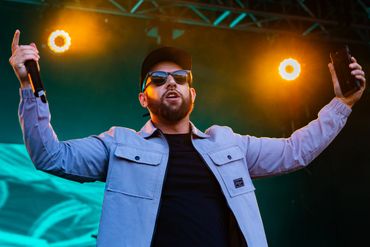 A bearded man wearing sunglasses performs on stage with a microphone and phone.