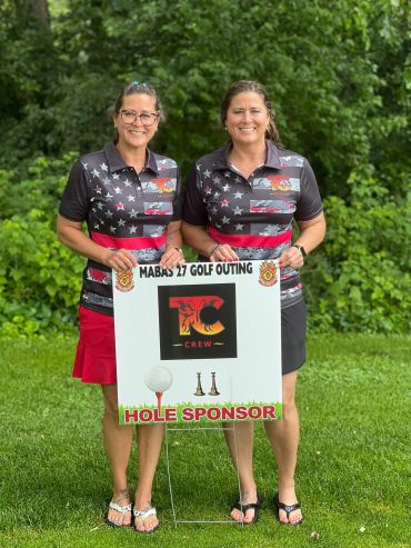 Two women posing with a golf outing hole sponsor sign.