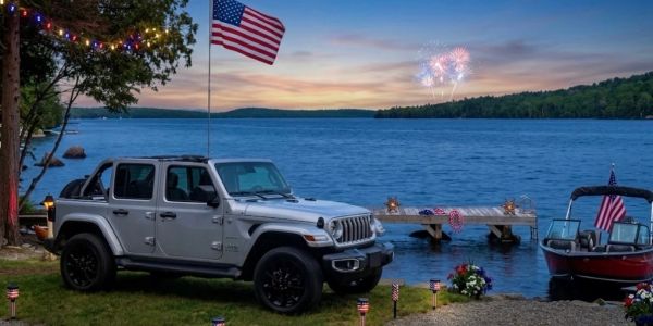 Silver Jeep parked by lakeside with American flag and fireworks.