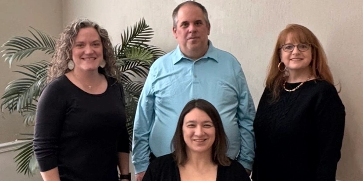 Four adults posing indoors with a plant in the background.