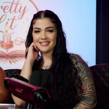 Smiling woman with long dark hair and tattoos, sitting in front of a microphone.