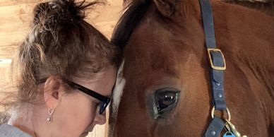 A woman gently touching foreheads with a brown horse inside a stable.