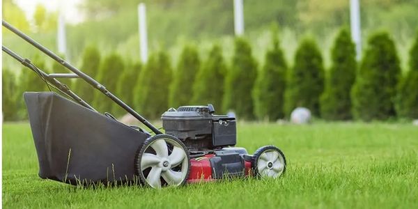 A lawn mower on green grass with a blurred background of trees and fence.
