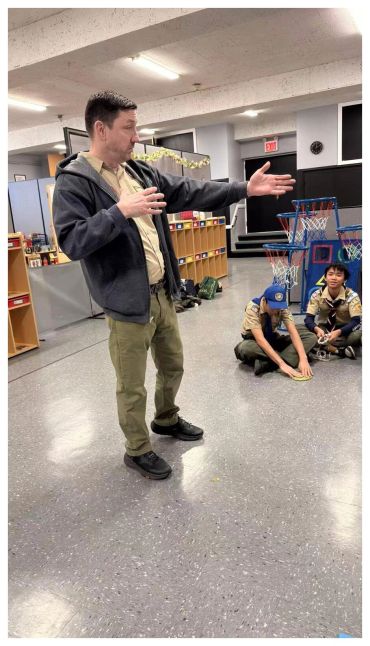 Man playfully posing while two boys in scout uniforms sit on the floor indoors.