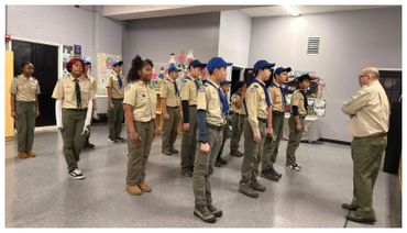 A group of scouts standing in formation indoors, listening to an adult leader.